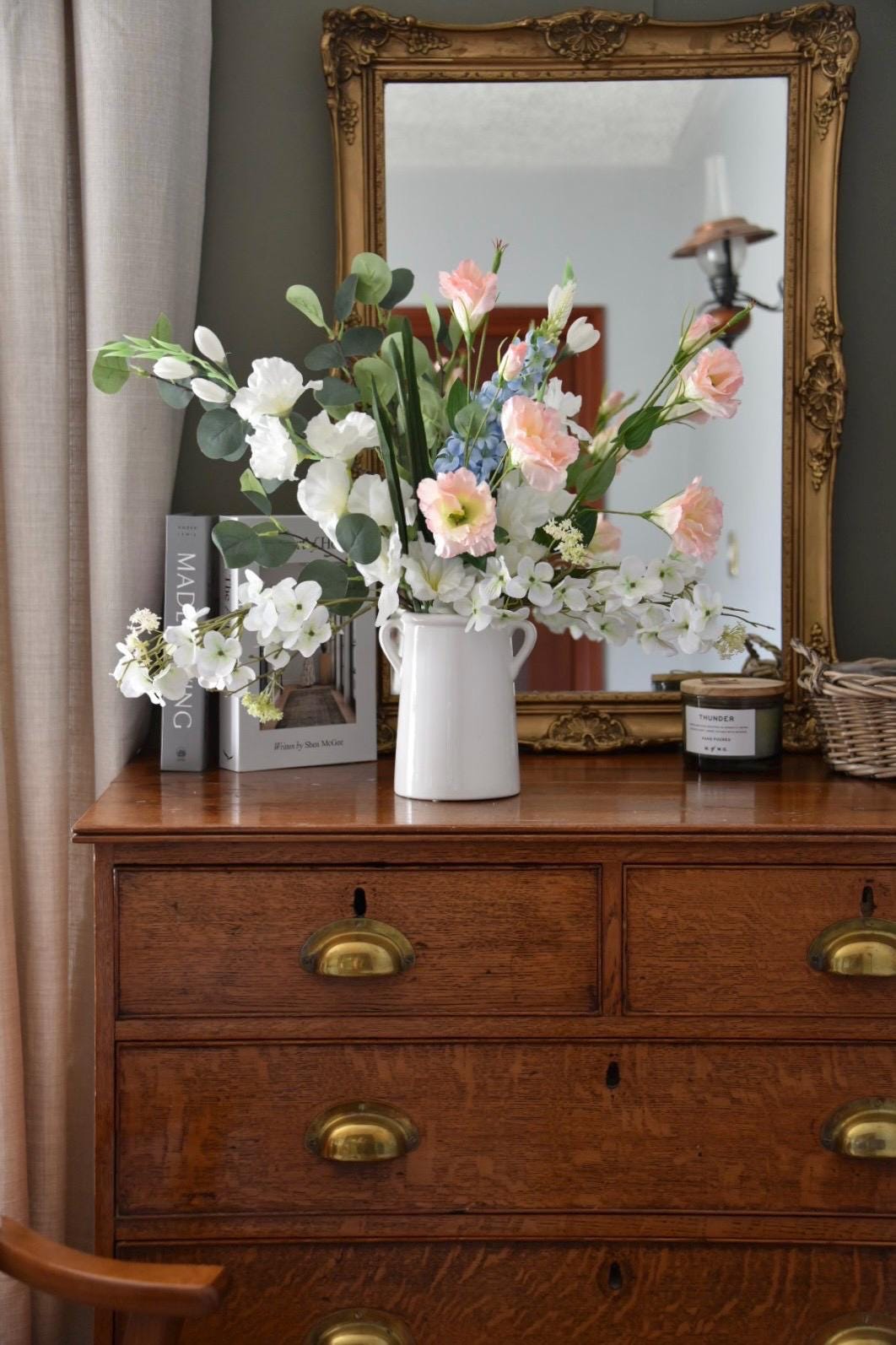 Wild flowers including gladiolus, wild white hydrangea, pink flowers, blue delphinium and green eucalytpus arranged in white vase on antique drawers