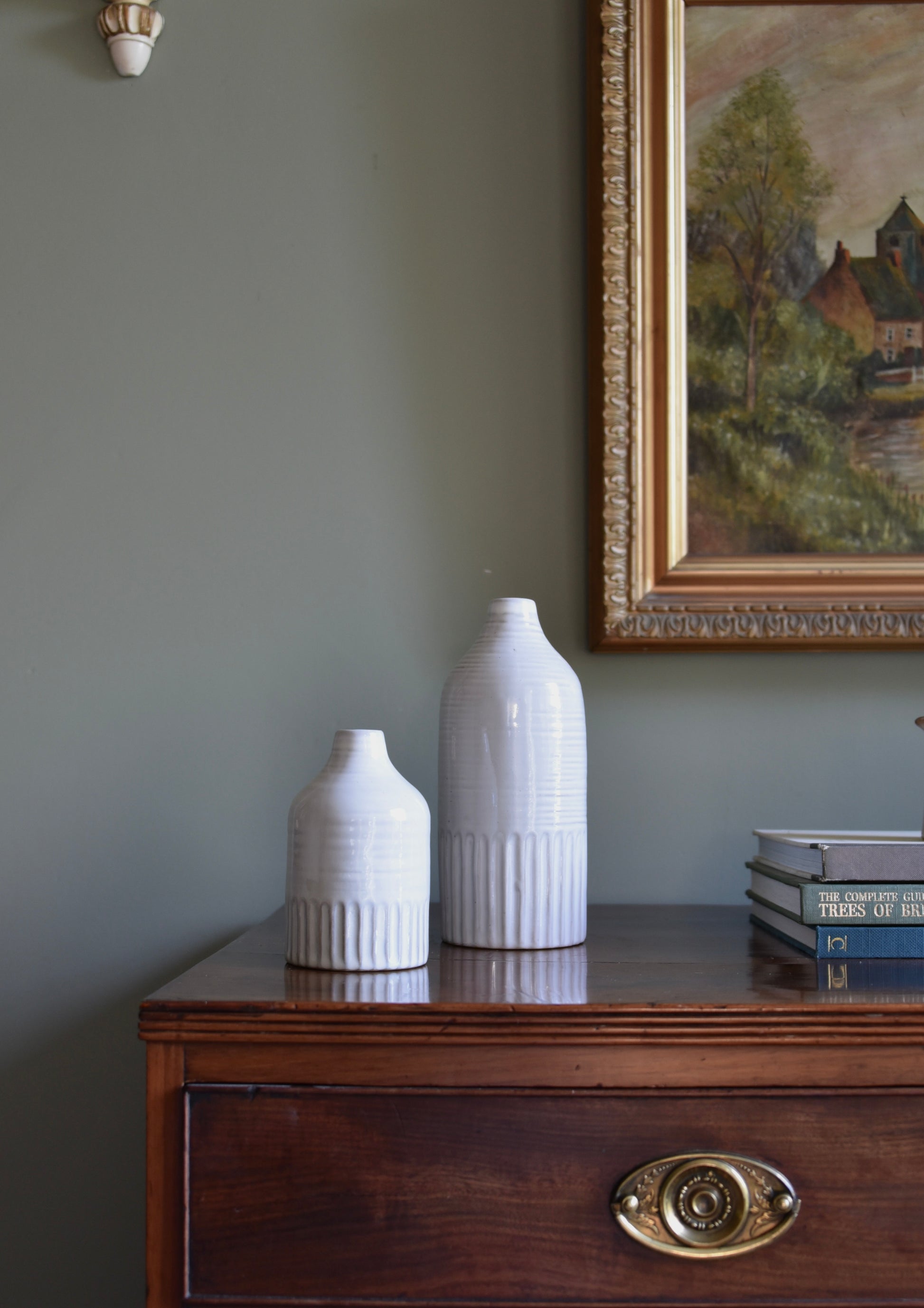 Two white ceramic bottles on a wooden dresser with an antique painting in the background.