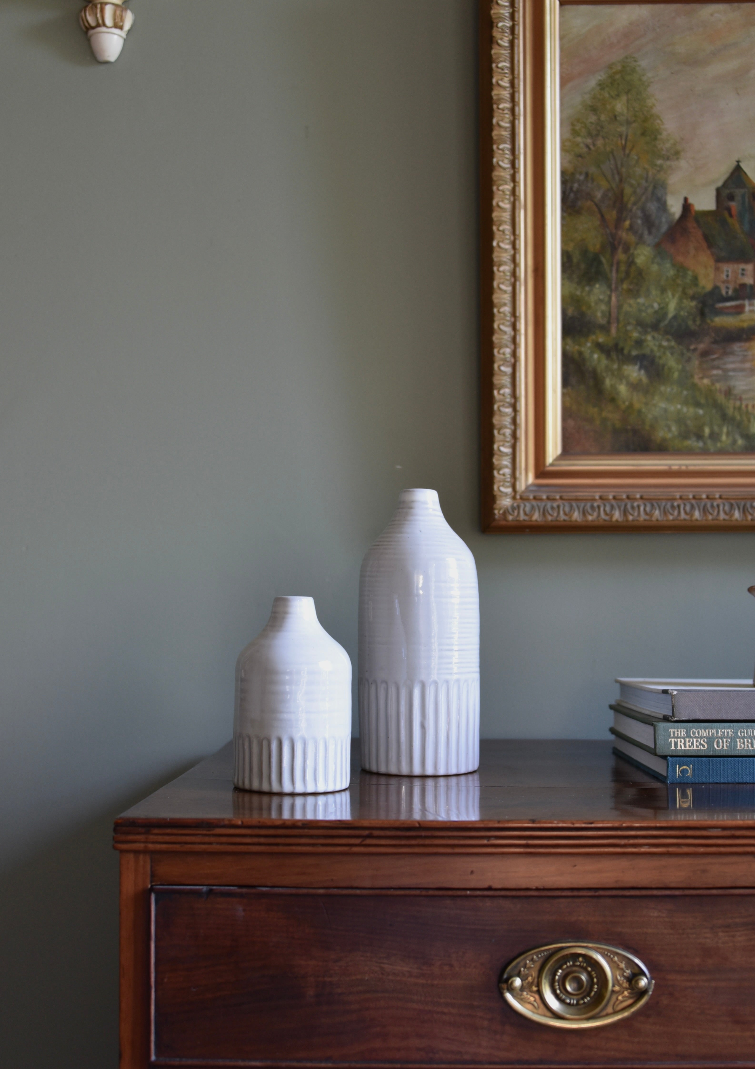 Two white ceramic bottles on a wooden dresser with an antique painting in the background.