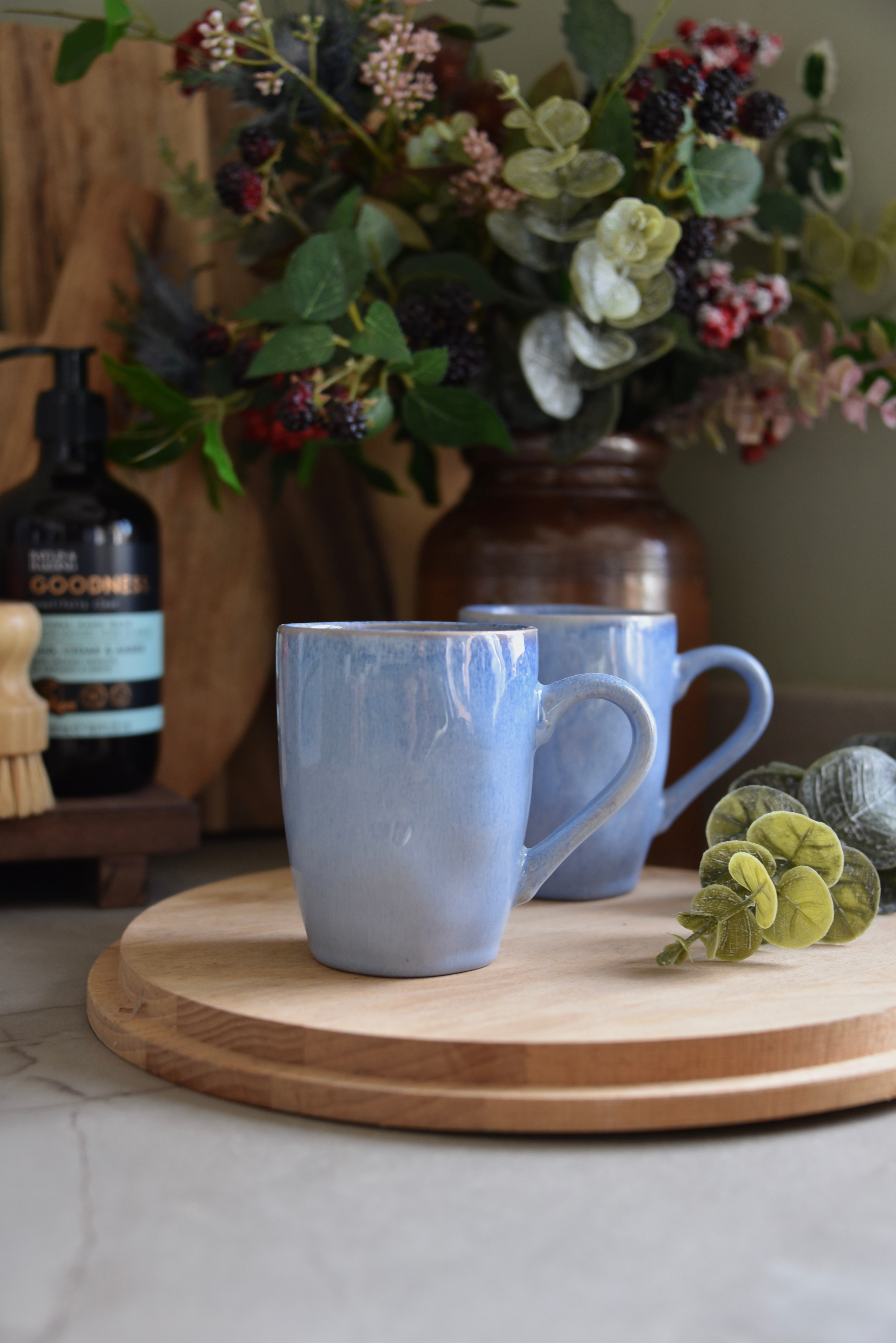 Two blue mugs on a wooden tray with flowers and a bottle in the background