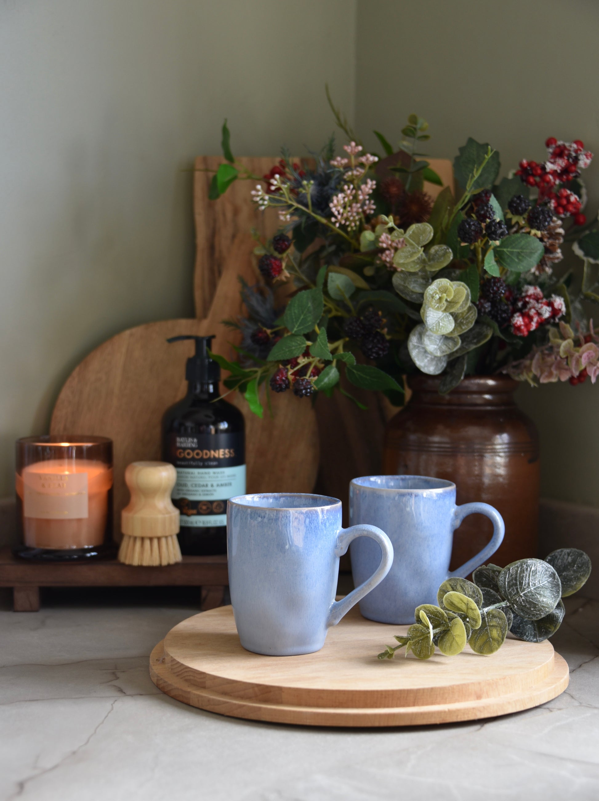 Two blue mugs on a wooden tray with a candle, brush, and decorative flowers in the background.