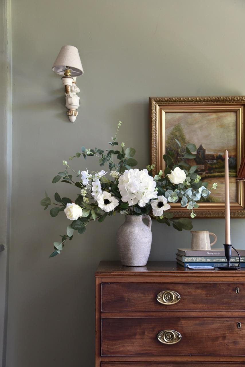 Faux flowers including white hydrangeas, cosmos, roses and wild eucalyptus styled on a wooden table and antique oil painting in the background. Sage green wall behind